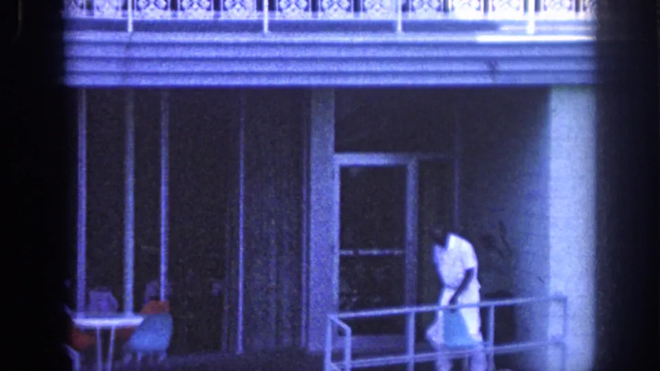 1963 Memphis frame showing a man in white clothing at the porch railing beneath a hotel balcony, with glass doors, columns, and shaded exterior walls behind him.