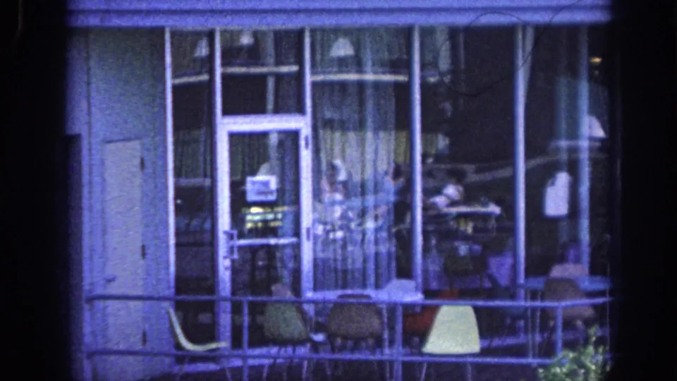 1963 Memphis 8mm frame of a lower balcony with a glass door, curtains, patio chairs, and a distant adult figure.