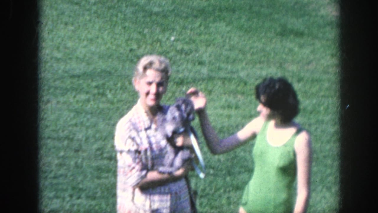 Two women stand on grass as one holds a small dog and the other reaches toward it.