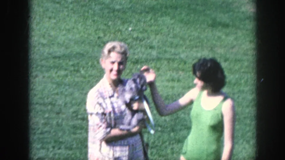 1963 Memphis home-movie frame showing two women on a lawn, one holding a small gray dog while the other reaches to pet it.