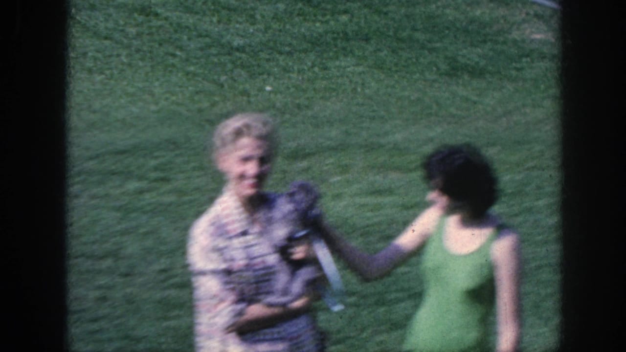 1963 Memphis frame with a woman holding a small dog as another woman in a green one-piece swimsuit reaches across on the lawn.