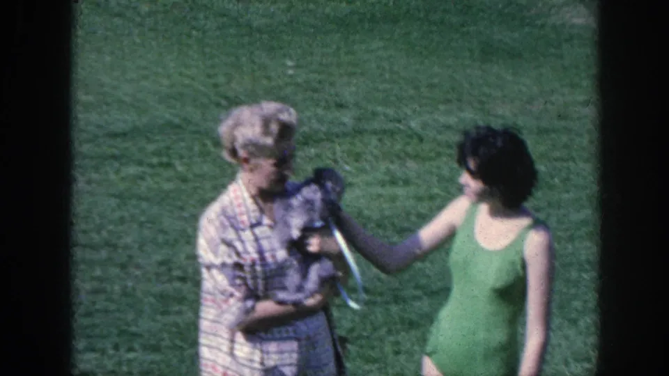 1963 Memphis home-movie still of a woman looking down at a small dog as another woman in green stands nearby on grass.