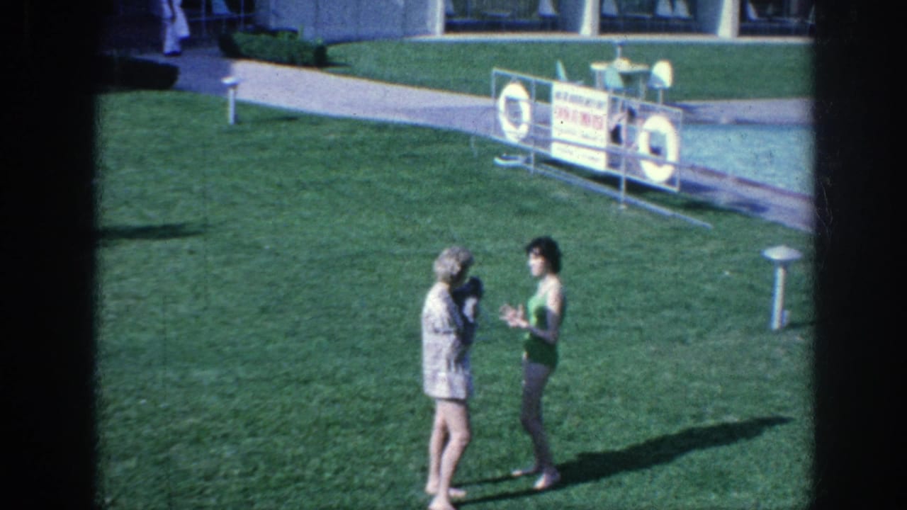 Two women face each other on a sunny lawn near a poolside sign.
