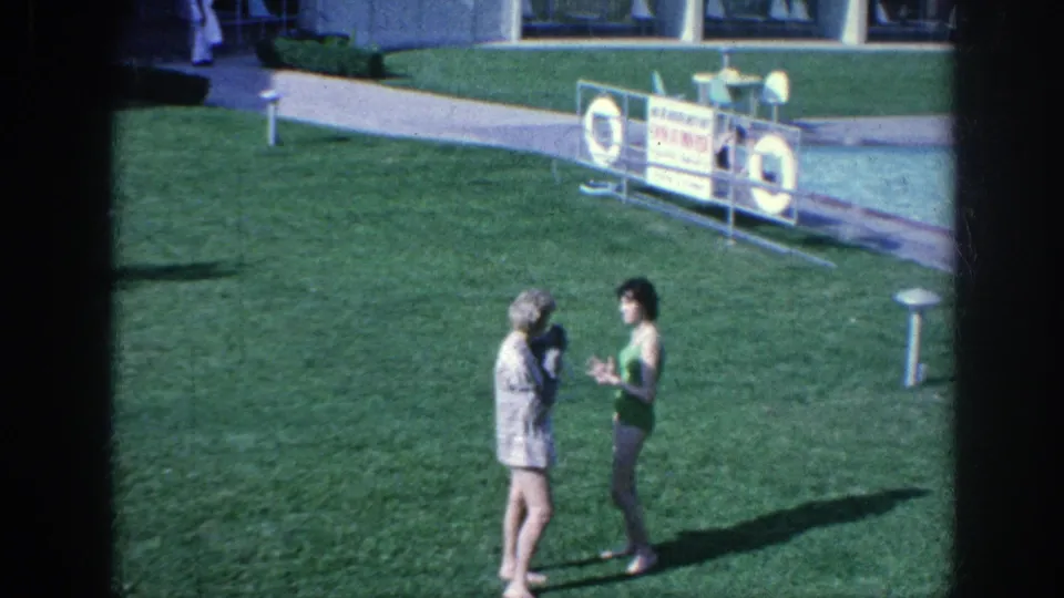 1963 Memphis-area poolside view with two women facing each other on the grass, a small poodle held at chest height, and safety signage behind them.