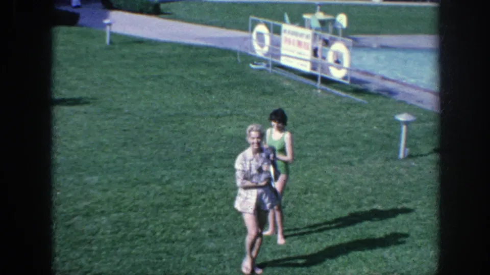 1963 Memphis-area 8mm frame of a woman walking across a poolside lawn holding a small poodle, with another woman in a swimsuit behind her.