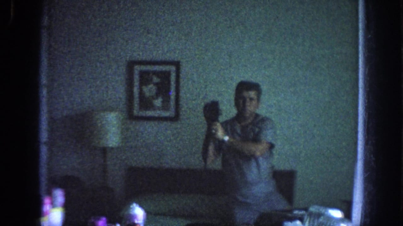 In 1963 Memphis home-movie footage, a man holds a camera toward a mirror in a dim interior with a lamp, sofa or bed edge, and framed wall art.