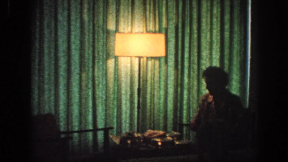 A silhouetted figure stands in a dim 1963 Memphis interior beside green curtains, a lit floor lamp, a chair, and a table with household objects.