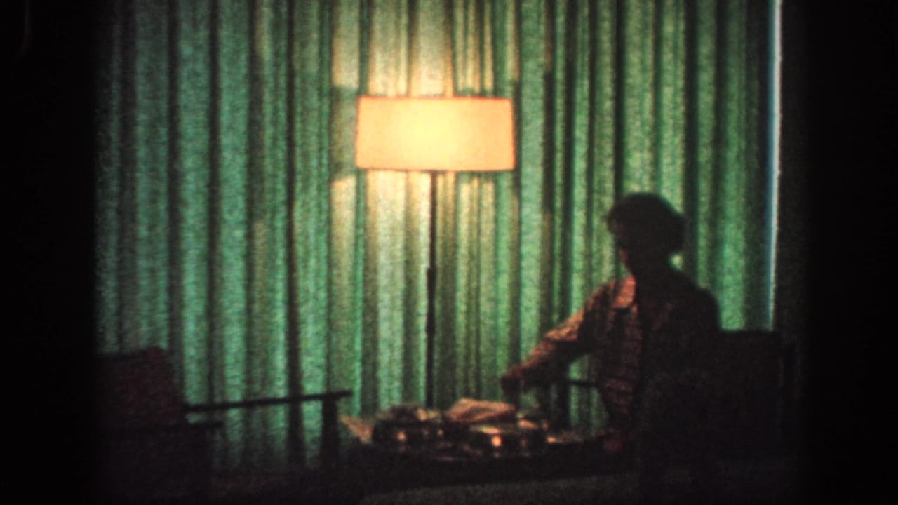 1963 Memphis 8mm frame showing a dim private room with green curtains, a glowing floor lamp, and an adult seated beside a low table.