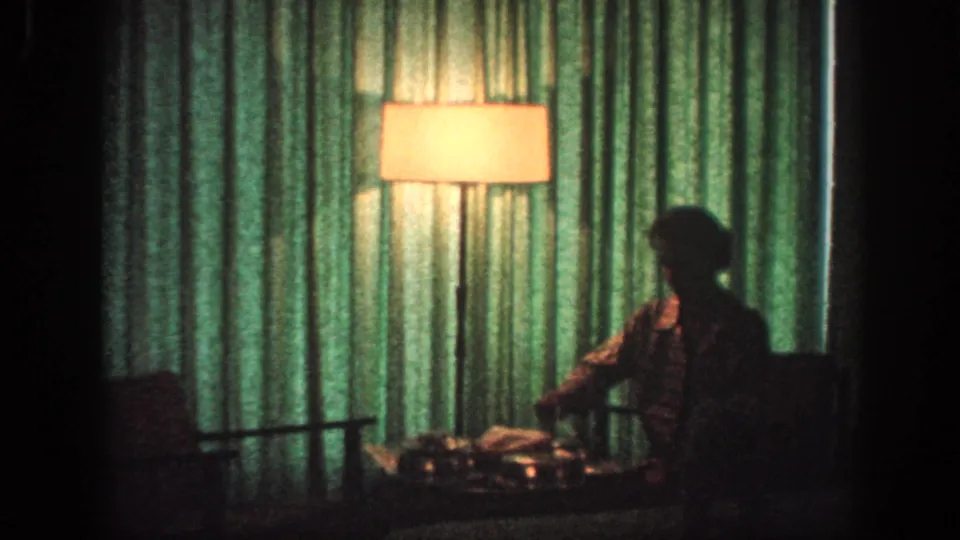 1963 Memphis 8mm frame showing a dim private room with green curtains, a glowing floor lamp, and an adult seated beside a low table.