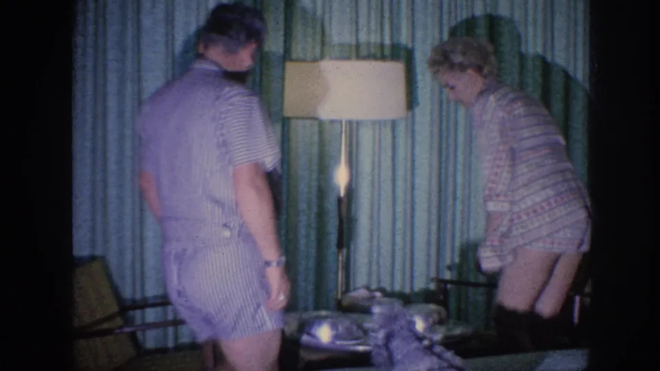 1963 Memphis 8mm still showing a man and woman in sleepwear standing near chairs, a lit floor lamp, and green curtains in a private interior.