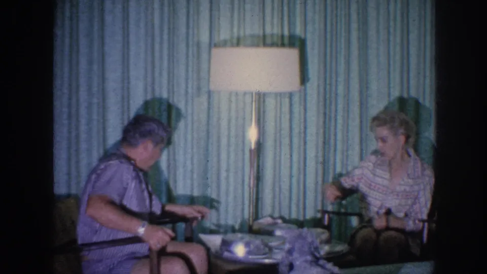 1963 Memphis home-movie frame of two adults seated near a small table, with a glowing floor lamp and green curtains behind them.