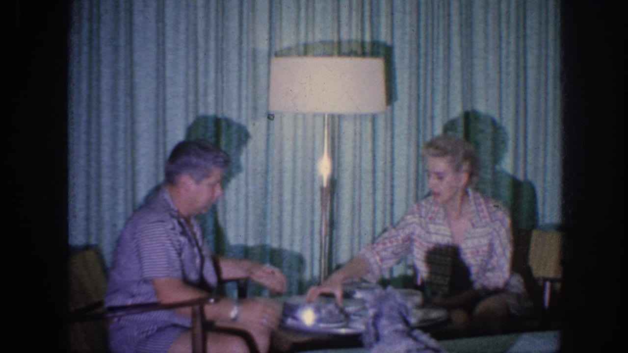 1963 Memphis home-movie frame of an elderly couple seated near plates and covered dishes under a tall lamp.