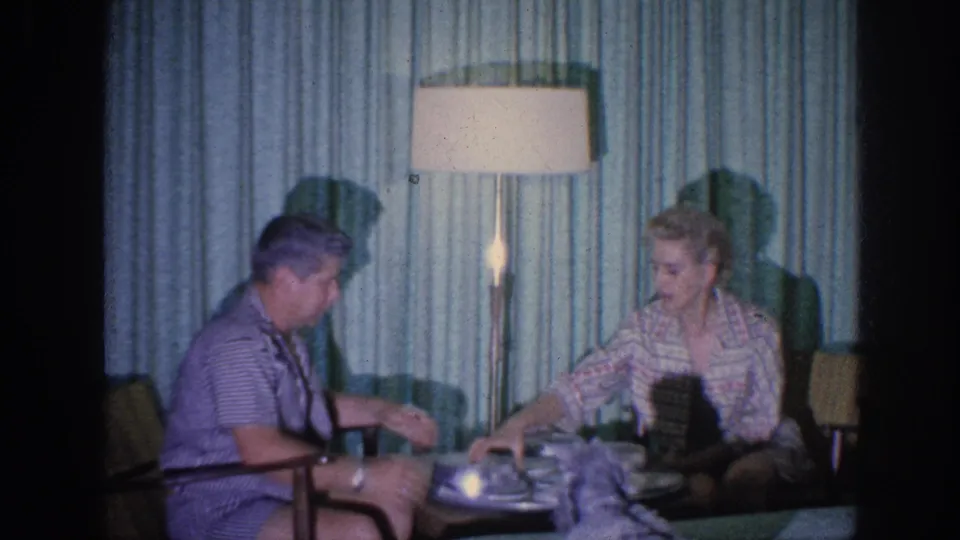 1963 Memphis home-movie frame of an elderly couple seated near plates and covered dishes under a tall lamp.