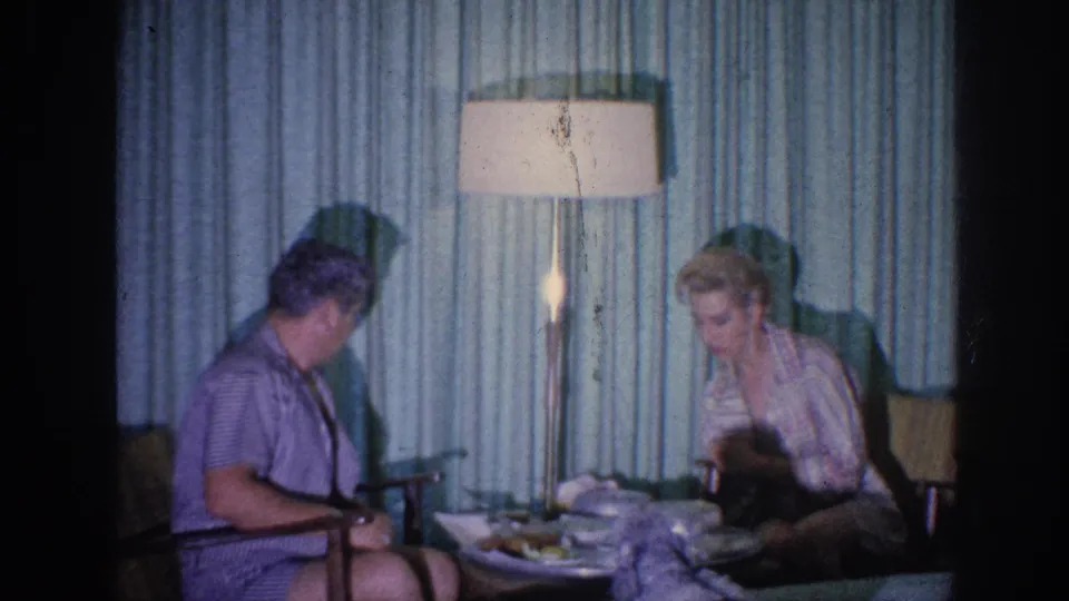 1963 Memphis home-movie still of an elderly couple at a meal, framed by blue-green curtains and a tall lamp.