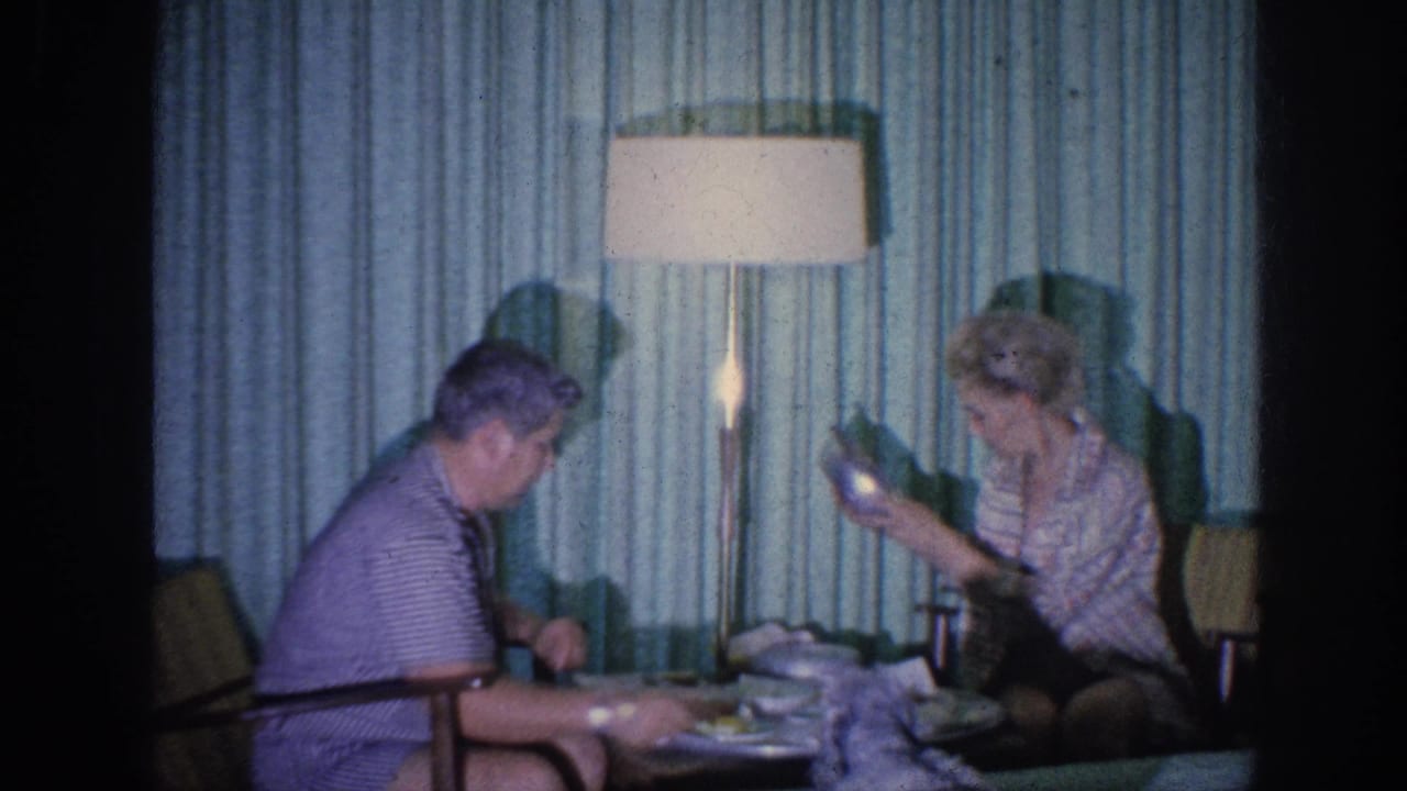 1963 Memphis home-movie frame of two adult women seated indoors near a low meal table, with teal curtains and a tall lamp in the background.