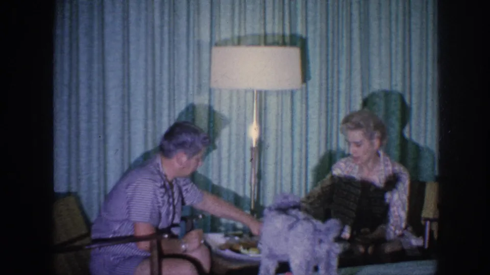 1963 Memphis home-movie frame showing a small dog reaching the low table between two adult women in a mid-century living room.