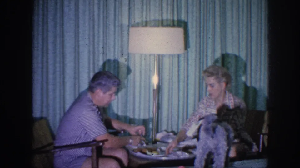 1963 Memphis home-movie frame of two adult women seated at a low meal table while a small dog stands in the foreground.