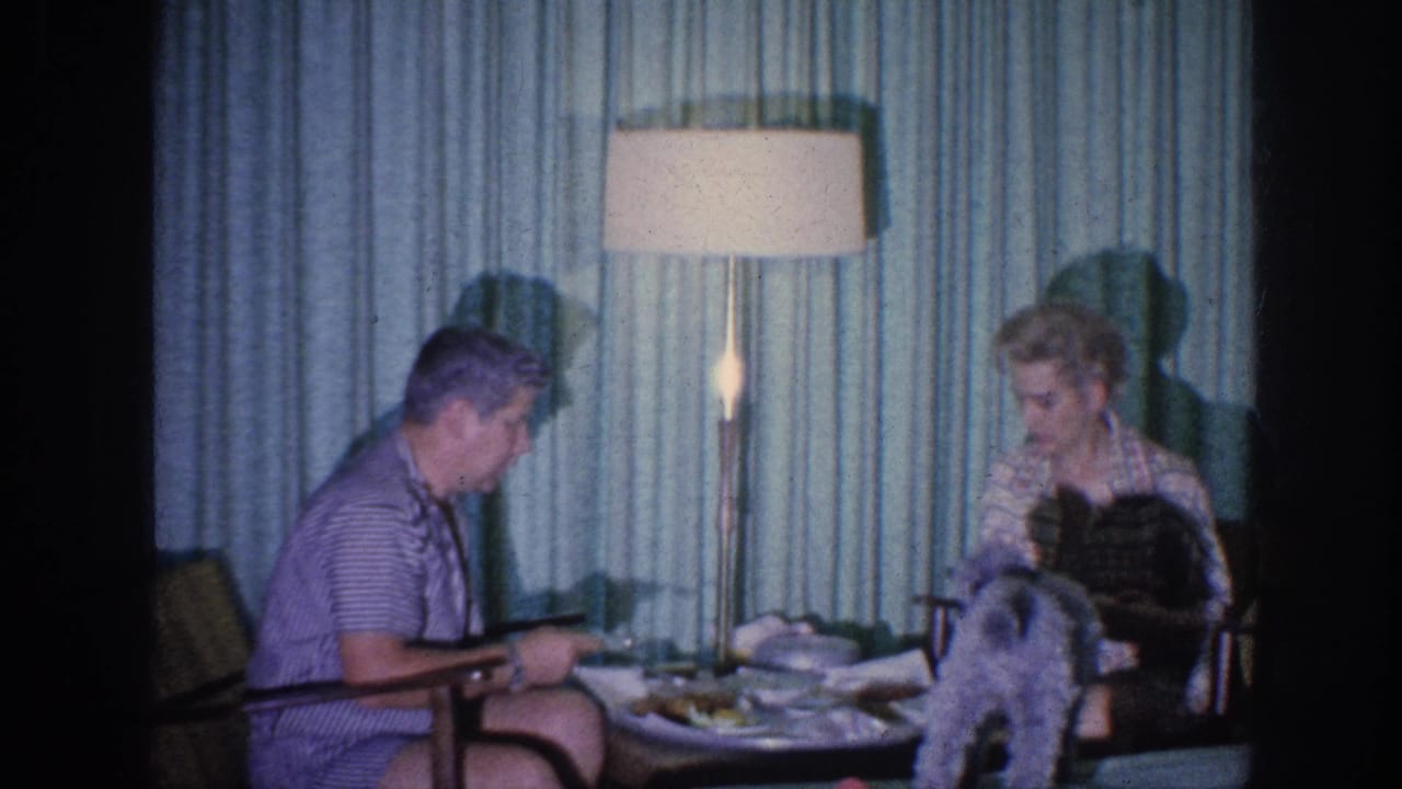 1963 Memphis home-movie frame showing a couple eating at a small hotel-room table under a floor lamp, with a small dog near the woman.