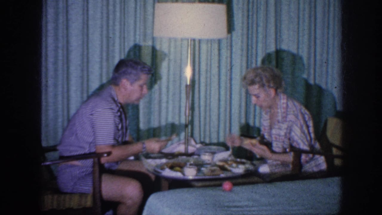Couple eating at a small table beside a bed, with curtains and a lit floor lamp behind them.
