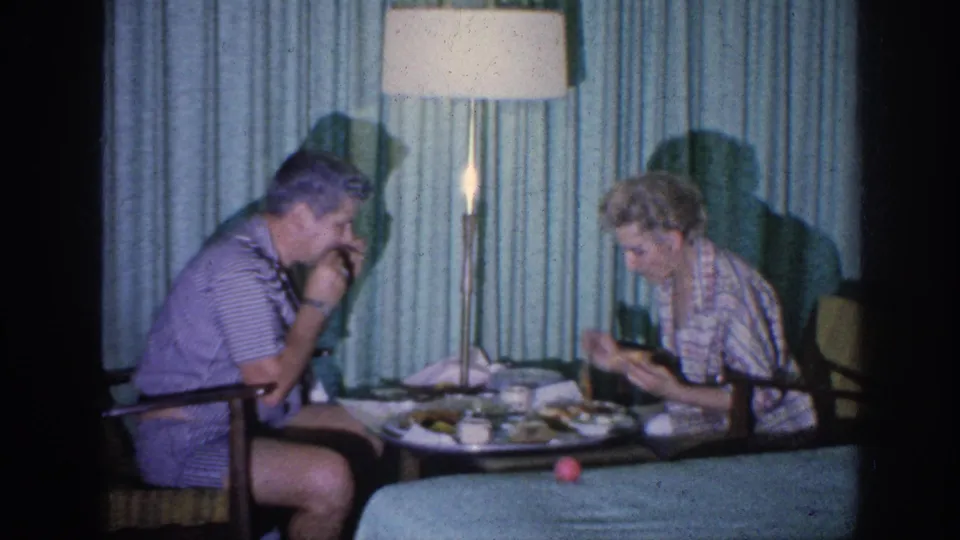 1963 Memphis home-movie frame of a couple eating quietly at a hotel-room table against blue-green curtains.