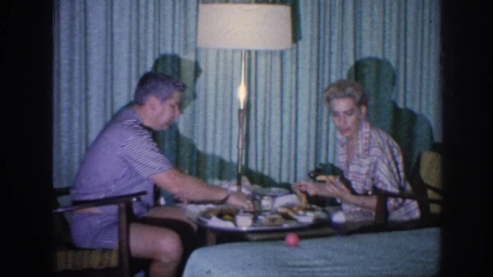 The man reaches toward the table as the woman looks forward with a cup during a 1963 Memphis living-room dinner scene.