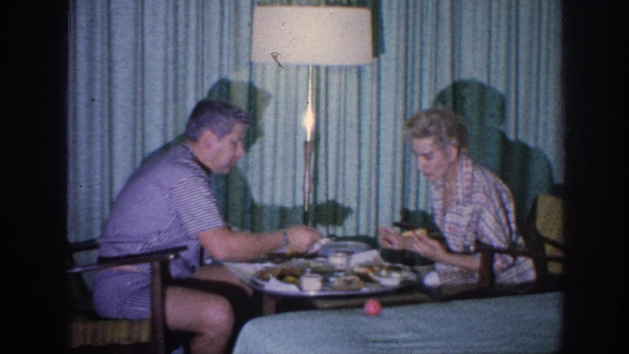In 1963 Memphis home-movie footage, a couple eats at a low table beside green curtains and a tall floor lamp.