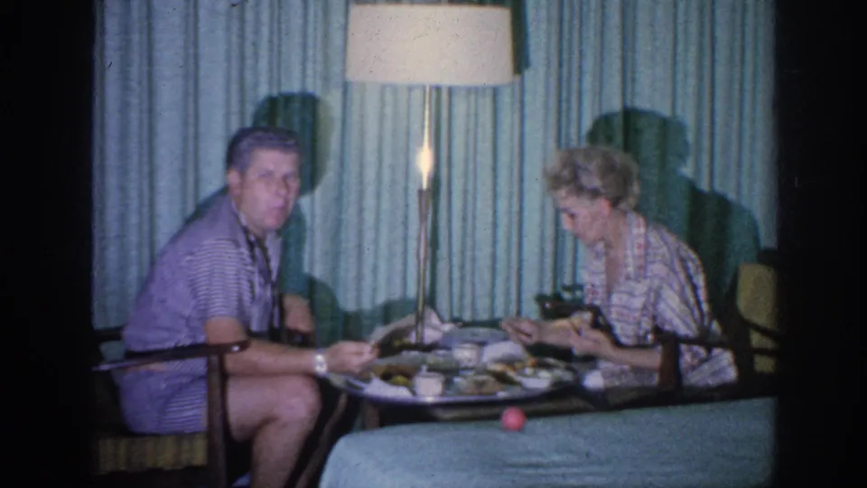 The man briefly looks outward from the dinner table as the woman remains focused on the meal in the 1963 room scene.