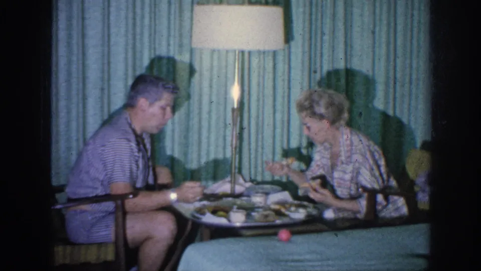 The couple remains seated at dinner in the Memphis interior, with food, cups, curtains, and a tall lamp visible.