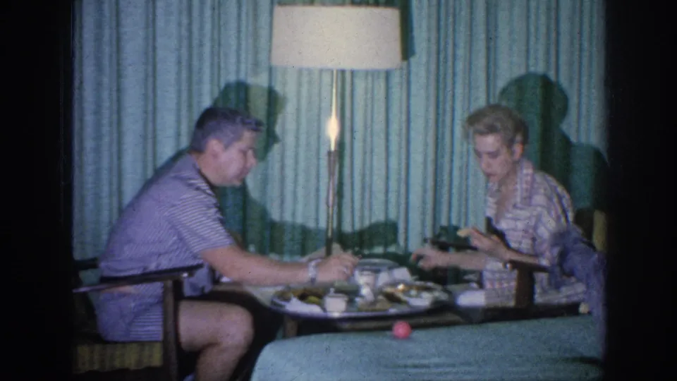 The man and woman continue eating from a tray in a Memphis hotel room, with plates, cups, curtains, and a lamp visible.