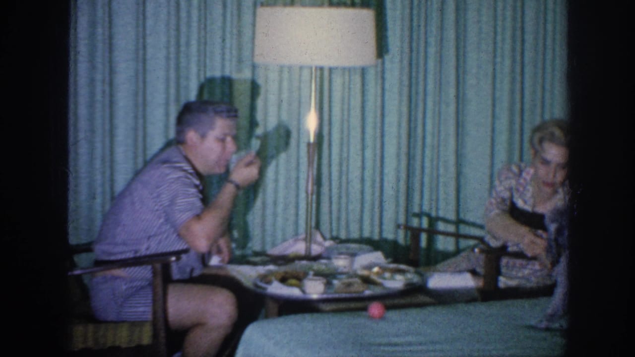 In a 1963 Memphis home-movie frame, two adults sit at a small meal table beside teal curtains and a lit floor lamp.