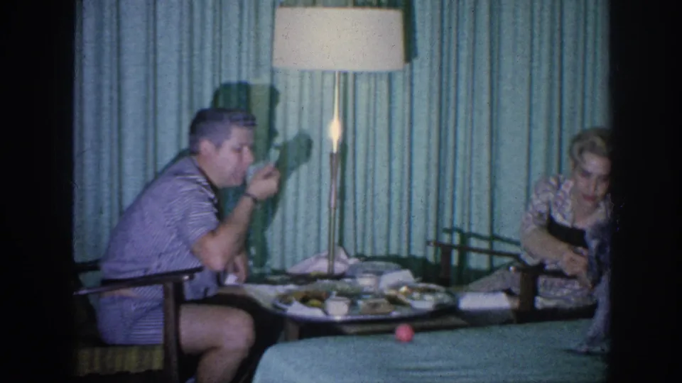 In a 1963 Memphis home-movie frame, two adults sit at a small meal table beside teal curtains and a lit floor lamp.