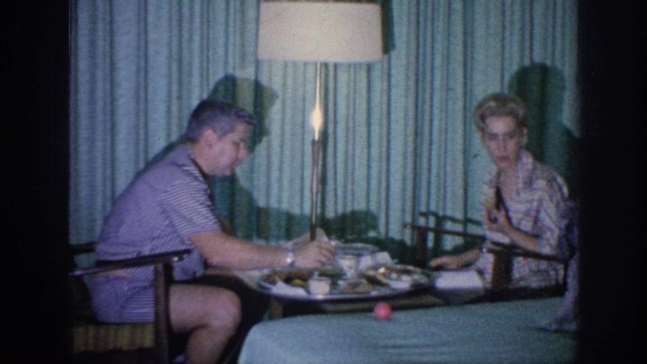 The man reaches across the meal table as the woman looks forward in the curtained Memphis interior.