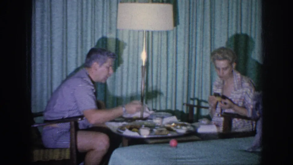A 1963 home-movie still shows two adults eating at a small table in a private Memphis room with teal curtains.