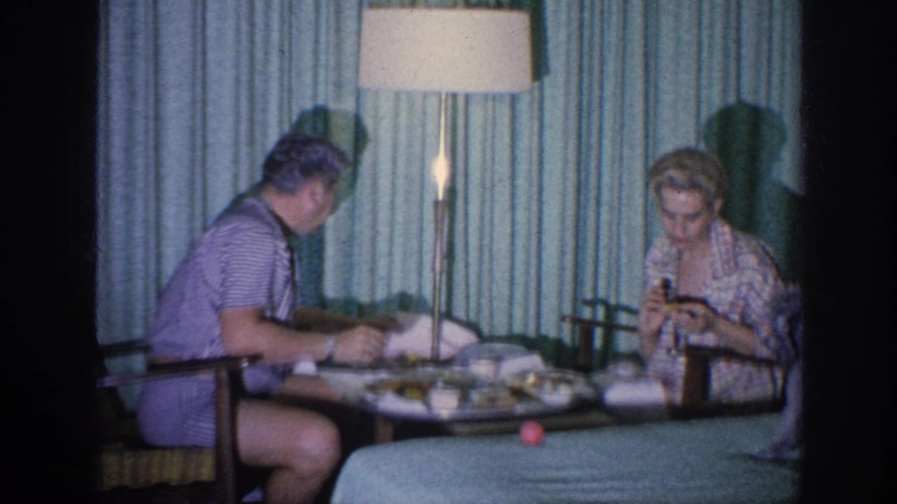 In a 1963 Memphis hotel-room home movie, a man and woman sit at a small table beneath a tall lamp with food and cups visible.