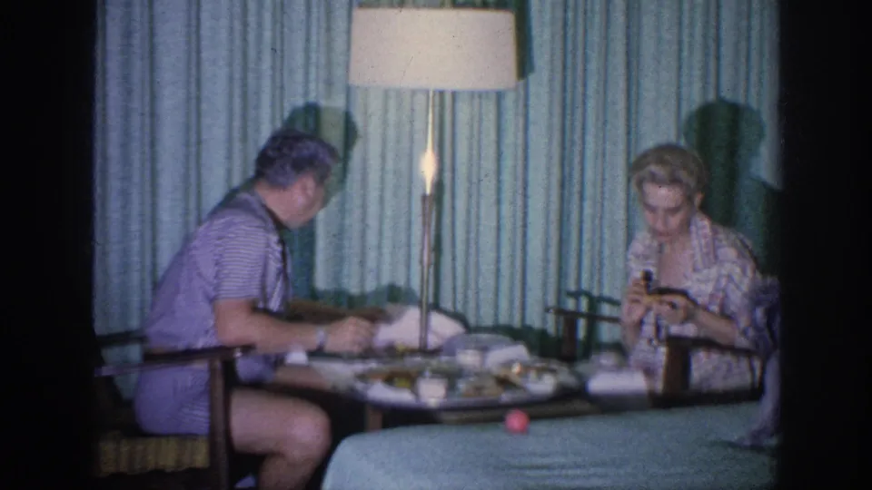 In a 1963 Memphis hotel-room home movie, a man and woman sit at a small table beneath a tall lamp with food and cups visible.