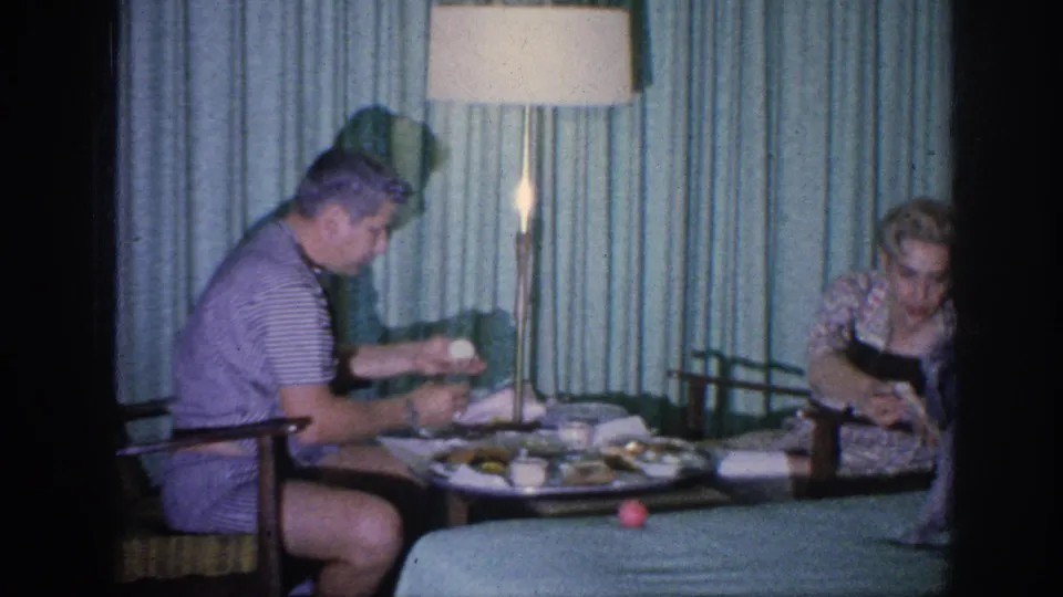 In the Memphis hotel room, the man holds food at the small table as the woman leans forward beside plates, cups, and a tall lamp.