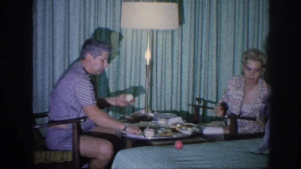 The final frame shows the couple at the hotel-room table, with the man turned toward the woman and food, cups, lamp, and curtains visible.