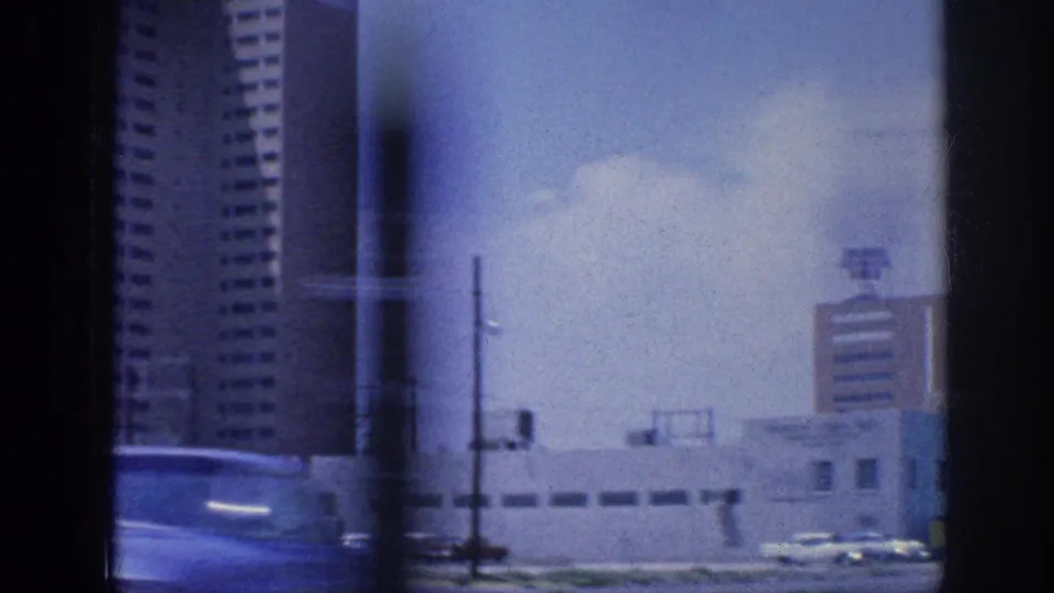 1963 Memphis travel footage showing a passing street view with tall buildings, utility poles, low rooftops, cars, and window reflections.