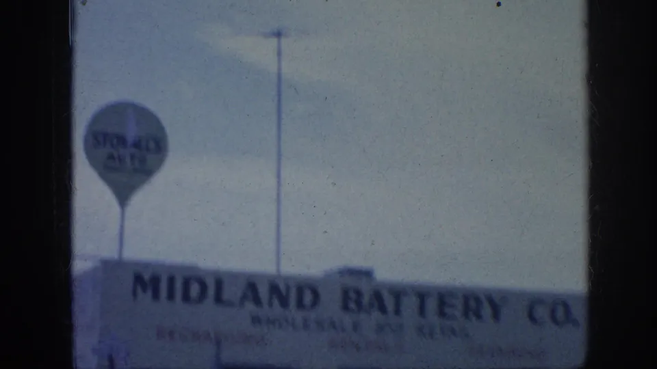 Closer 8mm view of Midland Battery Co. lettering with a round Stop gas sign above the roofline and partial street frontage below.