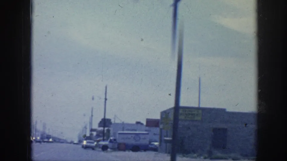 1963 Memphis industrial roadside seen from a moving vehicle, with utility poles, low buildings, cars, and a commercial truck near the right curb.