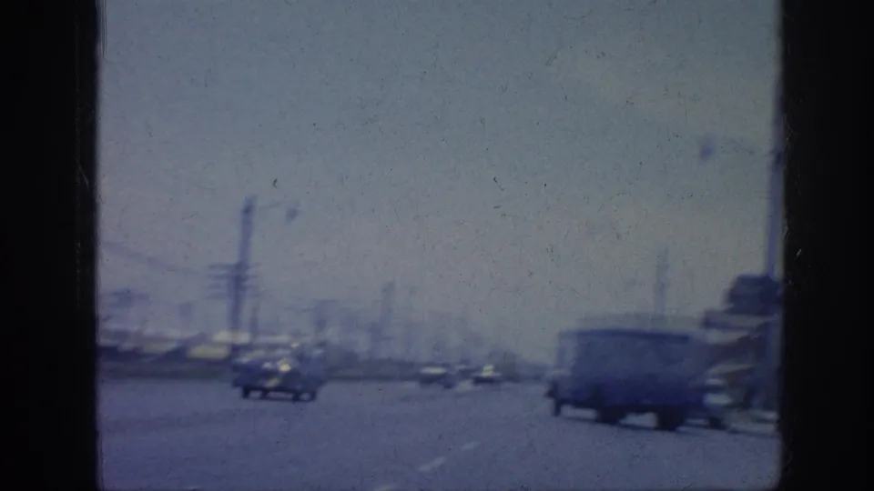 Moving-car 8mm view of a Memphis industrial road in 1963, with cars, a passing truck, utility poles, and motion-blurred buildings.