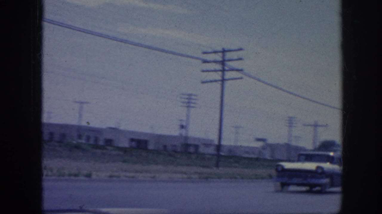 Roadside view from a moving vehicle with utility poles, wires, low buildings, and a passing car.