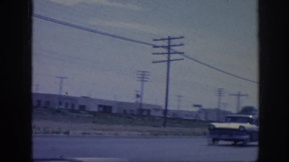 1963 8mm road-view frame with utility poles, overhead wires, low roadside buildings, and a mid-century car near the road.