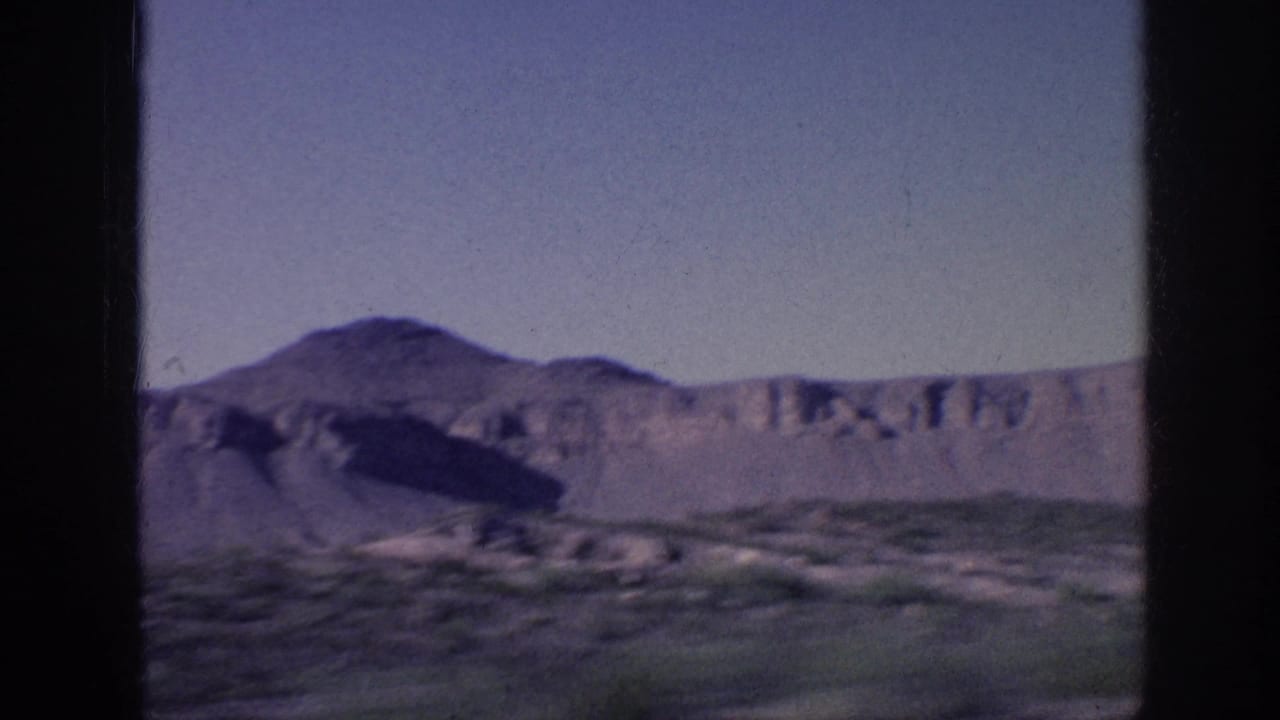Desert-like landscape with scrub foreground, pale cliffs, and a rounded mountain under open sky.