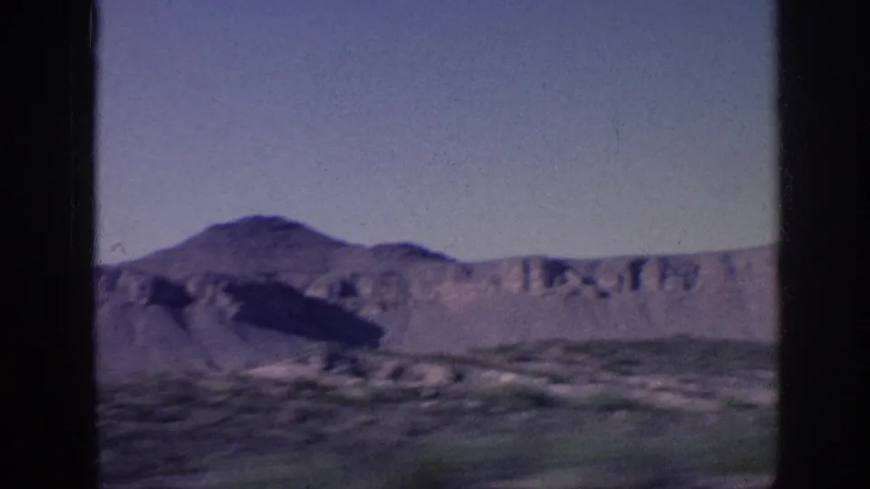 Home-movie travel frame showing desert scrub, pale cliffs, and a rounded mountain beneath a clear sky.
