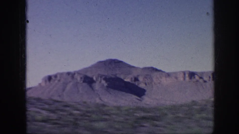 1960s 8mm frame of a broad scrub landscape with eroded cliffs and a low mountain ridge in soft faded color.