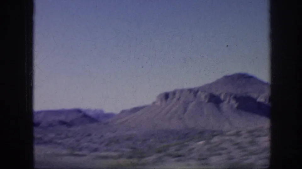 Faded 8mm travel frame showing open brushland, distant hills, and a mesa-like mountain form under clear sky.