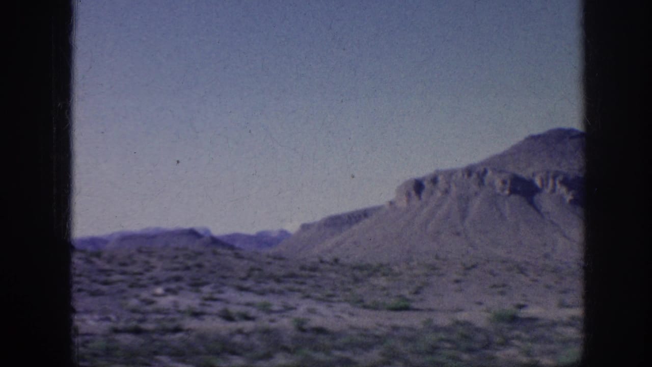 1963 8mm home-movie frame showing desert scrubland, sandy ground, and a low mountain under a pale sky from a moving travel viewpoint.