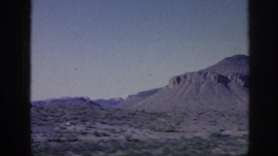 1963 8mm home-movie frame showing desert scrubland, sandy ground, and a low mountain under a pale sky from a moving travel viewpoint.