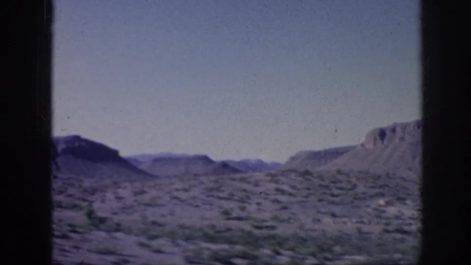 1963 8mm travel frame with open arid terrain, scattered scrub, distant hills, and dark vertical edges from the camera or vehicle window.